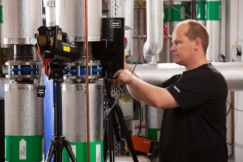 Kiwa technician calibrating industrial equipment in a facility, using a RiggersPro device and tripod, with insulated pipes and control panels in the background