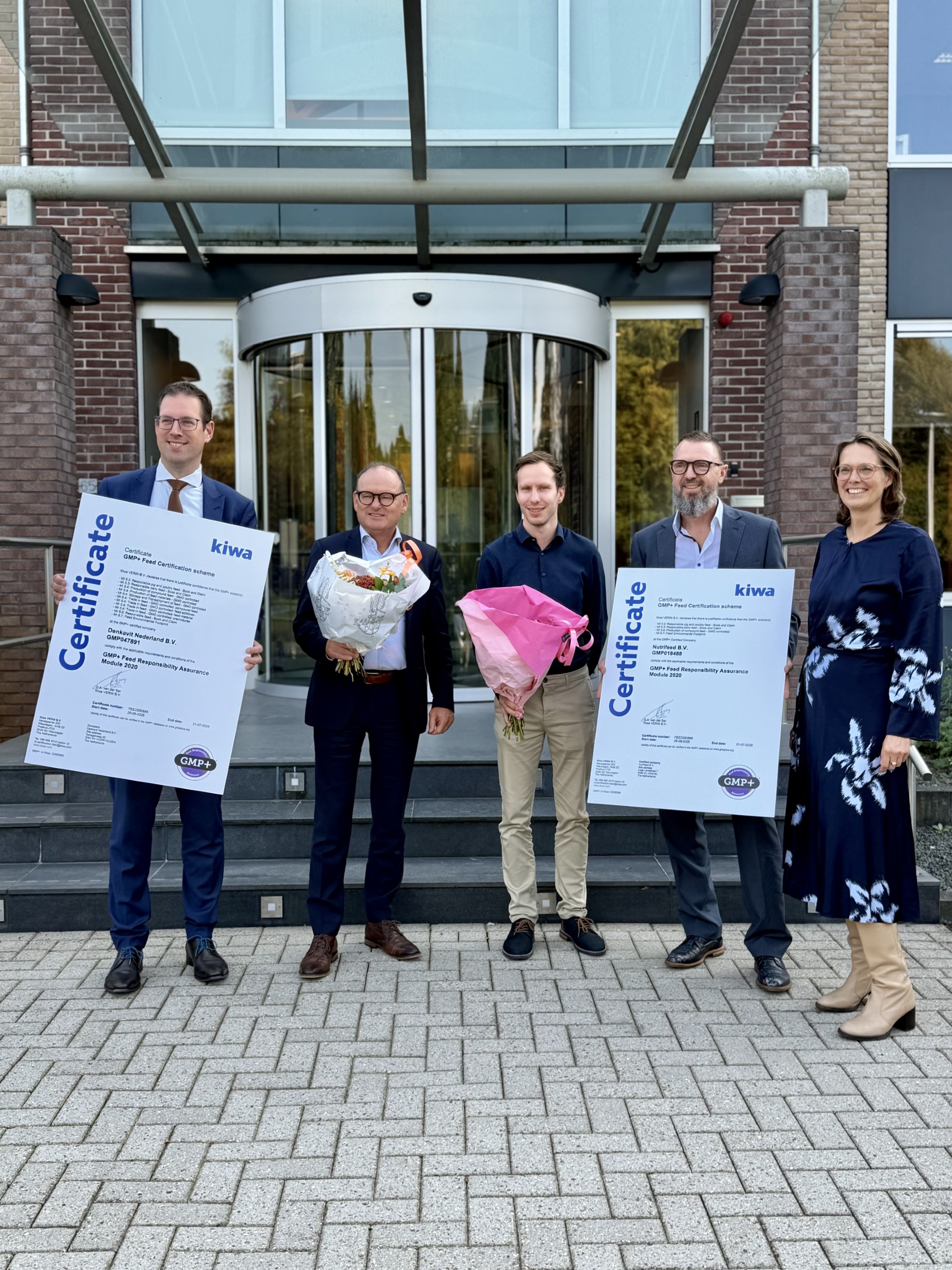 Five people standing outside an office building, holding certificates and flower bouquets for a GMP+ food certification presentation.