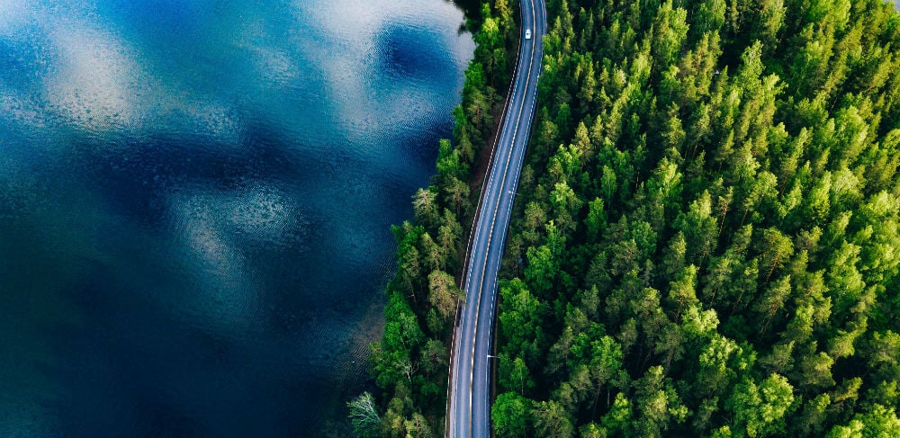 Bilden är tagen från ovan och visar en väg som går längst med en sjö och genom ett skogslandskap.