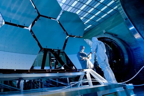 Scientists in clean suits working on large hexagonal mirrors in a laboratory, highlighting advanced technology and precision engineering