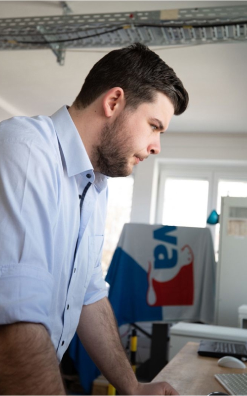 Kiwa lab employee in a laboratory setting focusing intently on a computer screen, wearing a light blue shirt; Lab filled with technical equipment