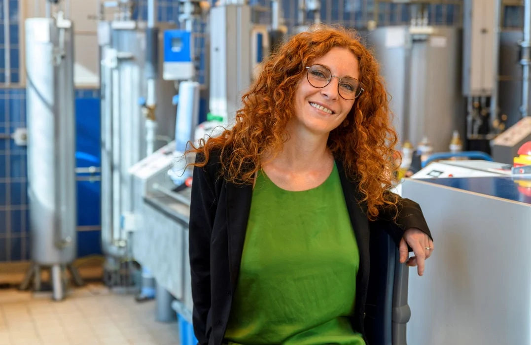 Giulia Botta, Global Business Development Manager for Energy Transition, with curly red hair and glasses standing in a modern laboratory, wearing a green blouse and black blazer