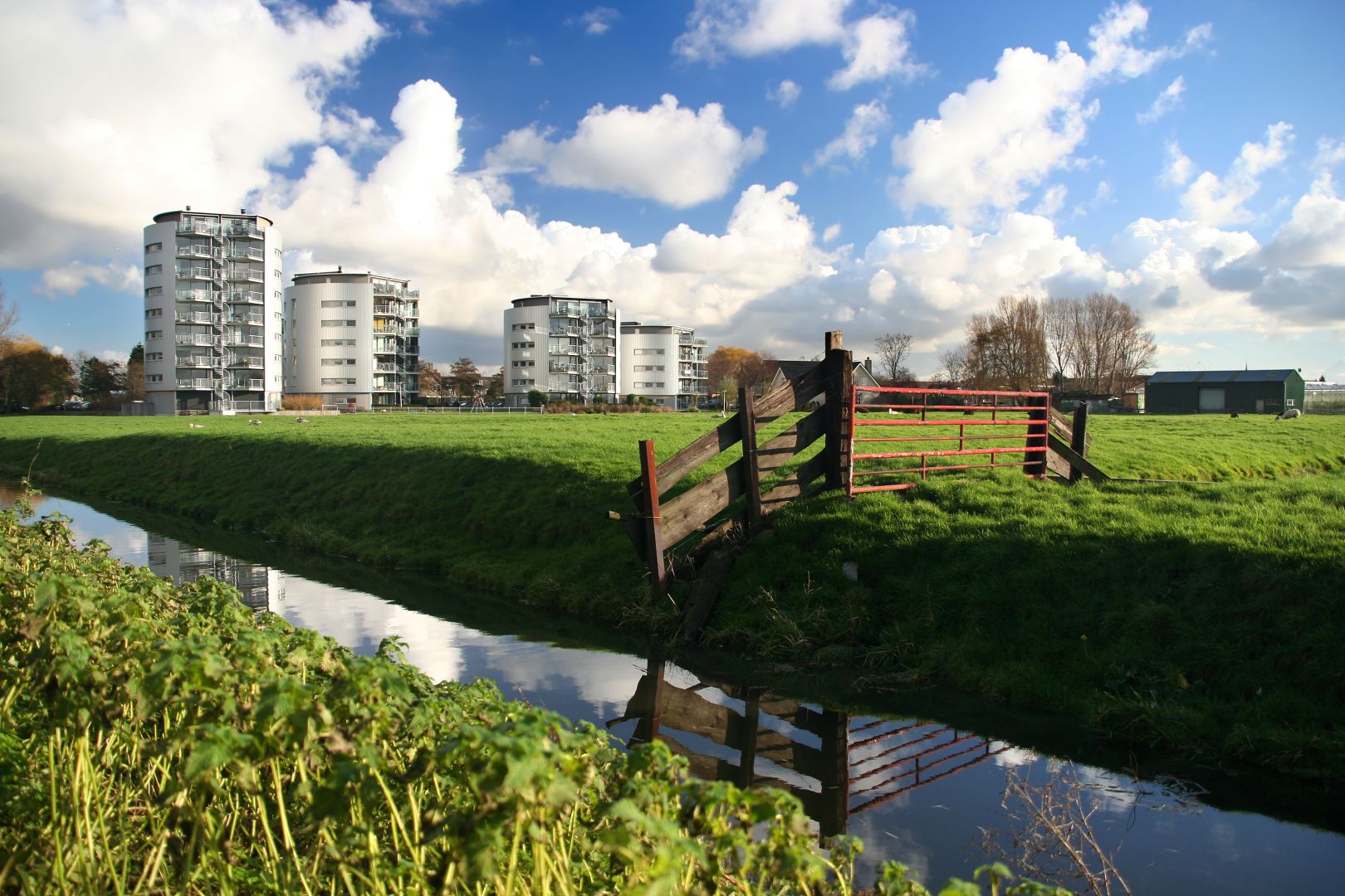 Grassy riverside landscape with modern apartment buildings, a red gate, and a canal under a cloudy blue sky.