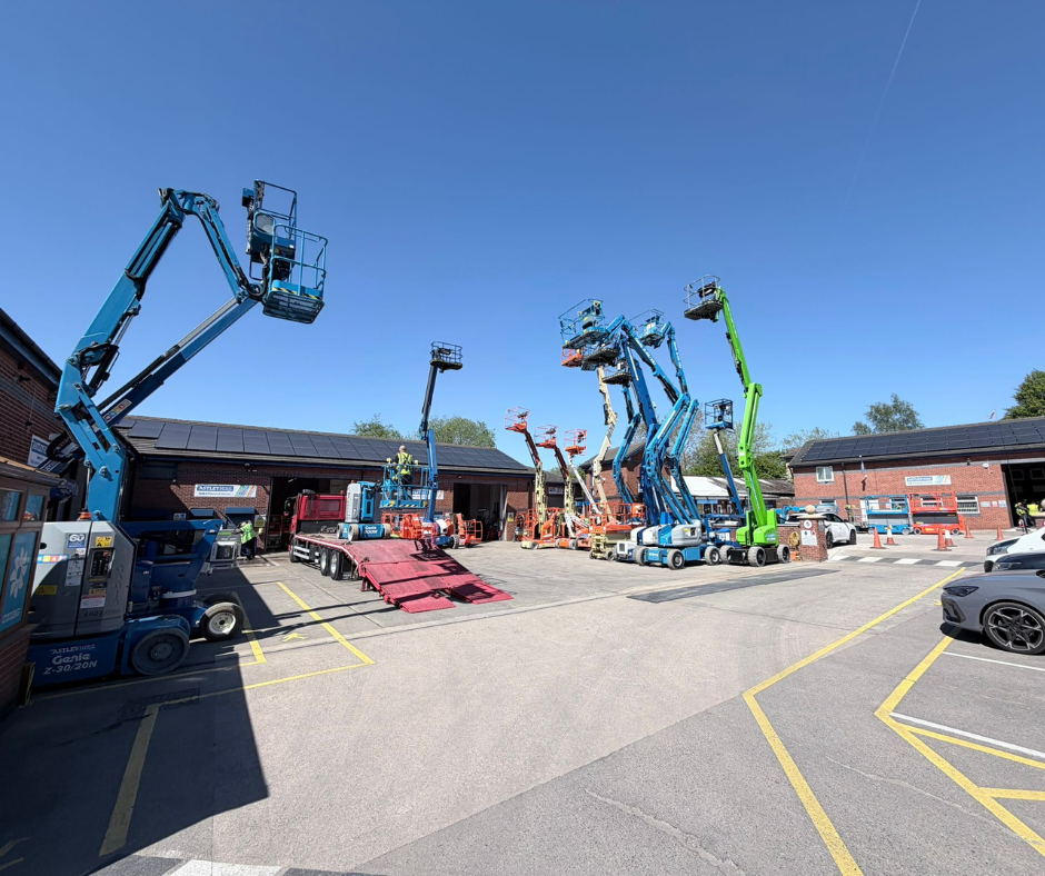 Row of elevated boom lifts and cherry pickers parked in a lot outside brick industrial buildings under a clear blue sky.
