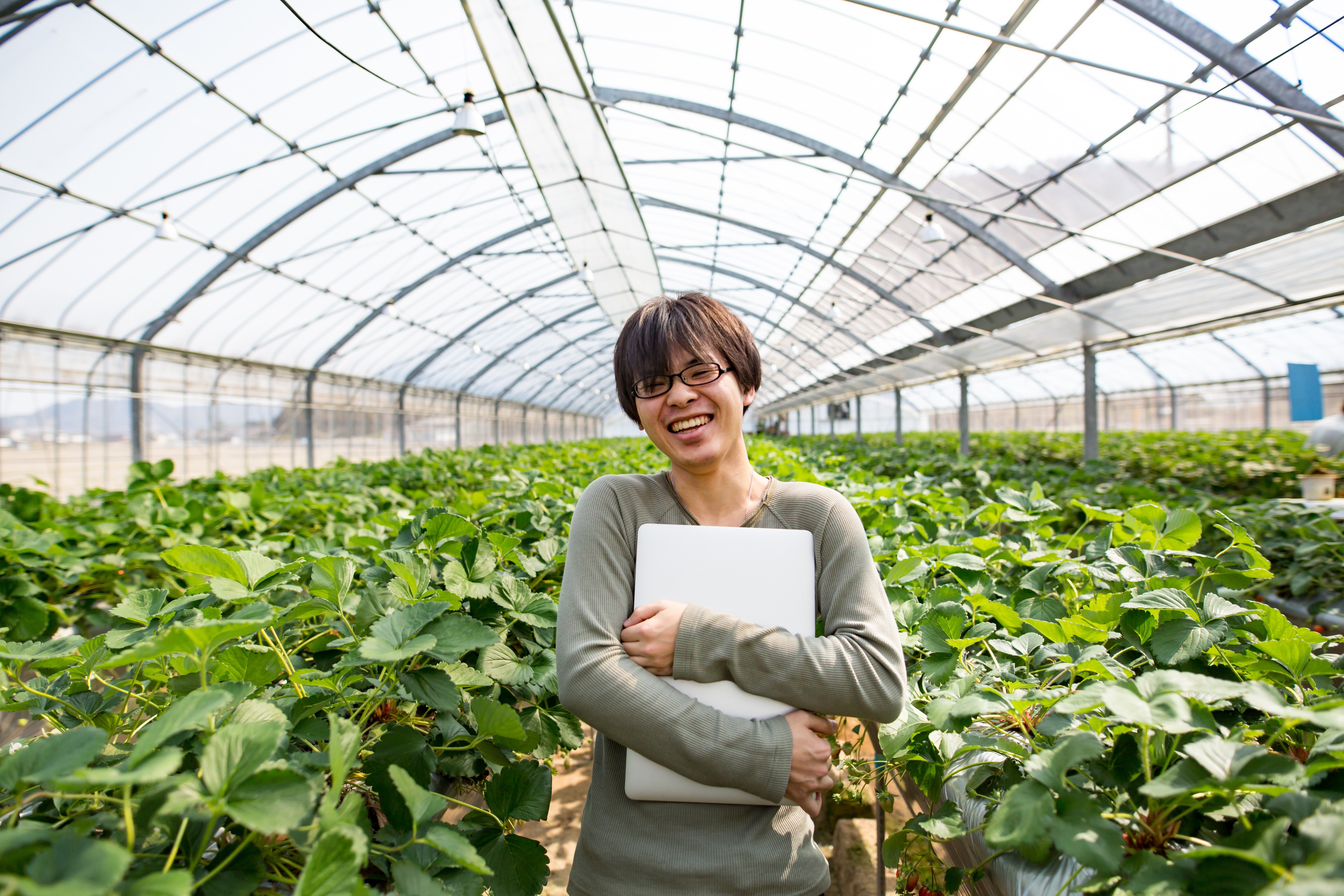 Portrait of a smiling woman holding a laptop in a greenhouse filled with green plants.