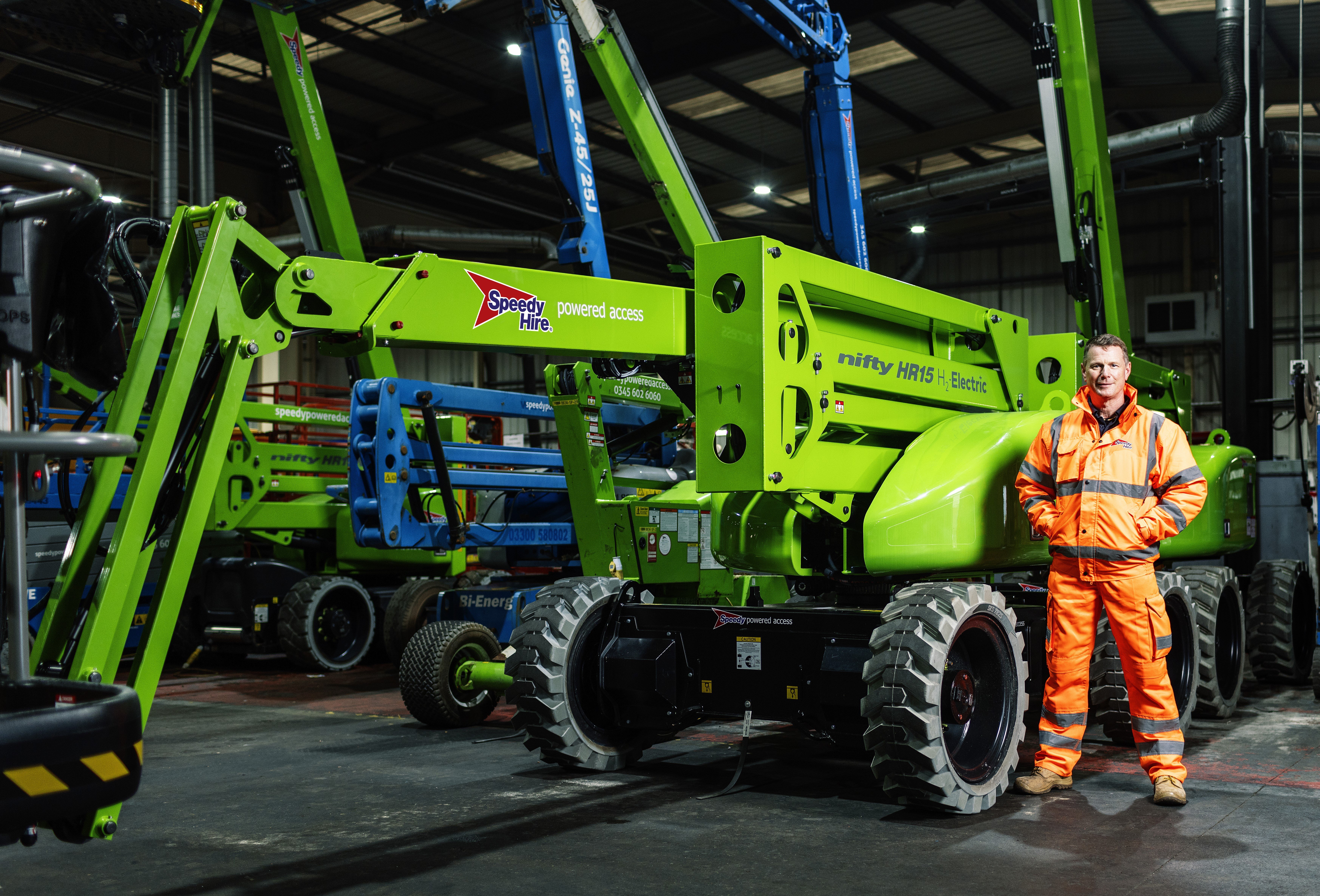 Man in orange high-visibility workwear standing beside a lime green Genie HR15 Hybrid electric boom lift in an indoor equipment warehouse.