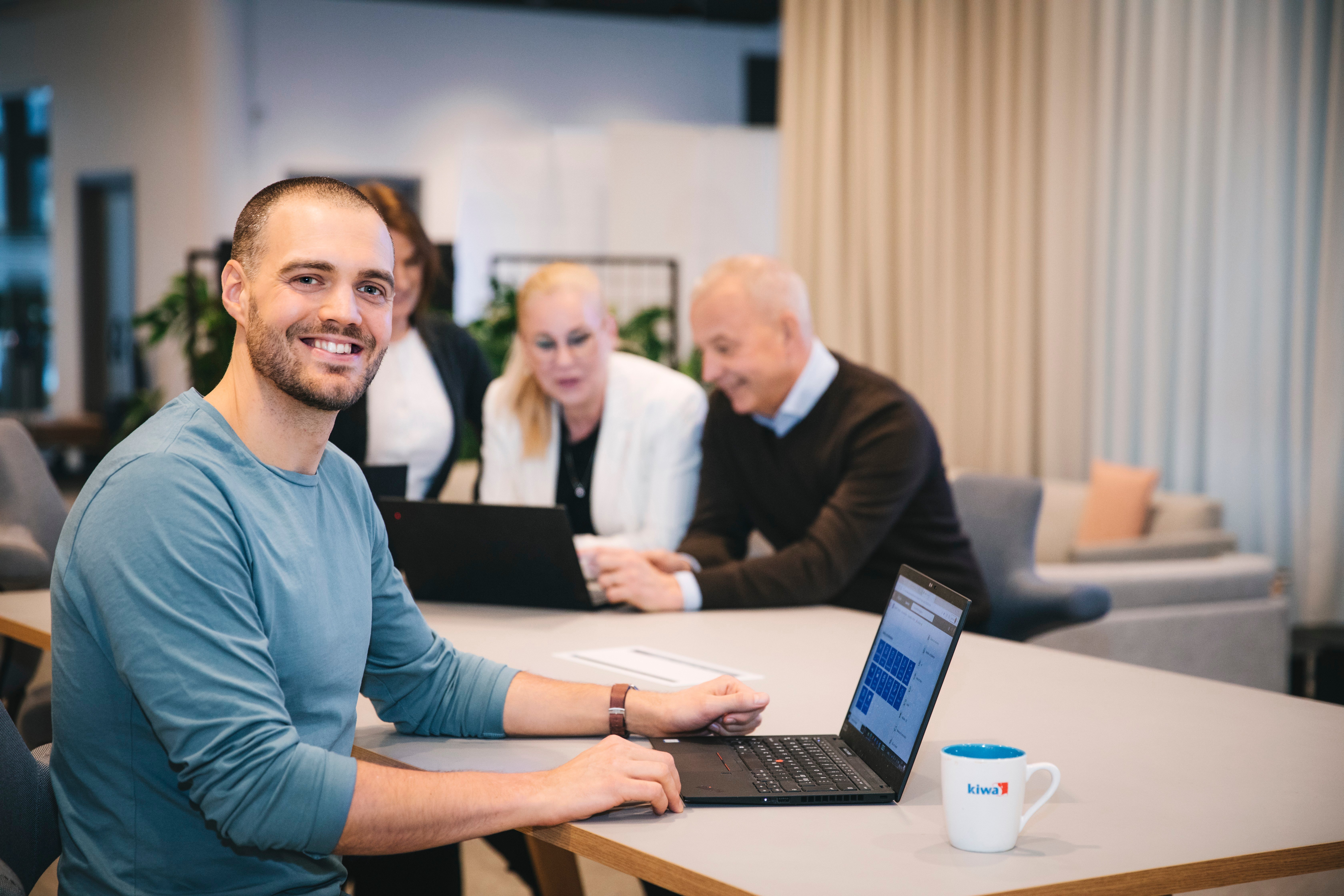 Man sitter vid laptop i kontorsmiljö med kollegor i bakgrunden. Affärsmöte med dator och kaffekopp på bordet.