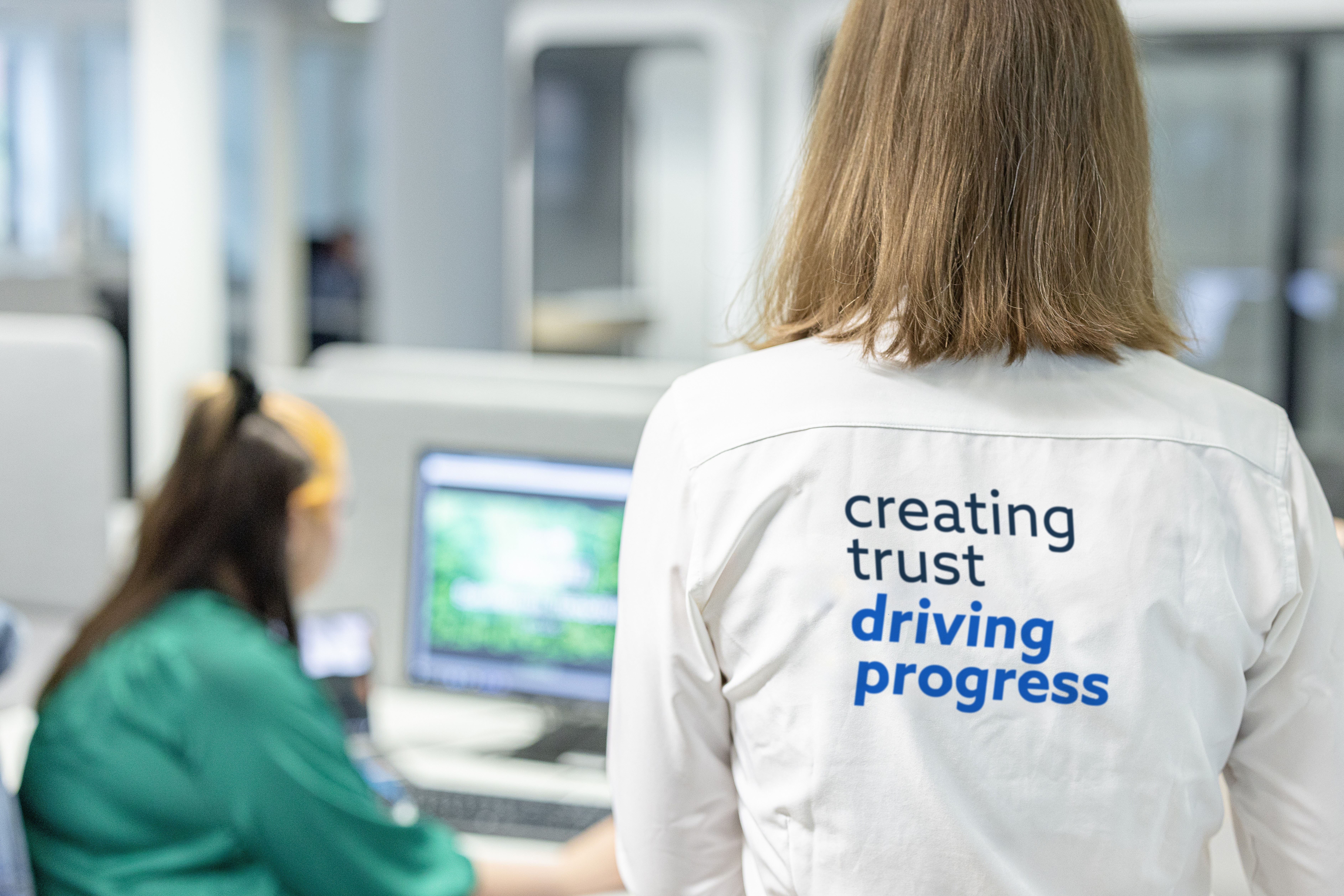 Person i vit skjorta med texten ”creating trust driving progress” i kontorsmiljö, med en kollega vid datorn i bakgrunden.