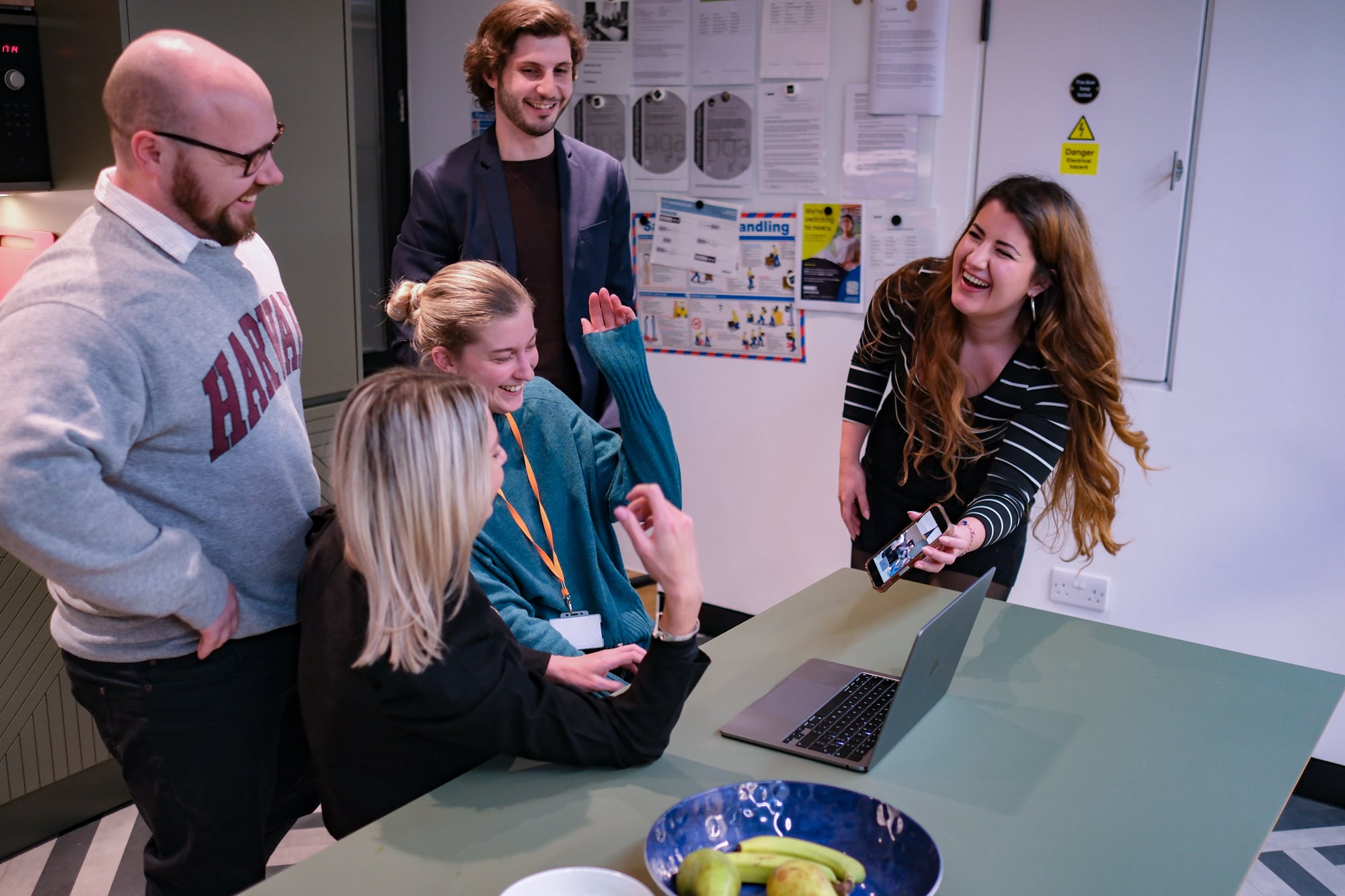 Group of five coworkers gathered around a laptop in an office, smiling and chatting while one woman holds up a smartphone.
