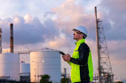 Industrial engineer in high-visibility clothing and hard hat operates equipment at an oil refinery with storage tanks and smokestacks in the background during a cloudy sunset