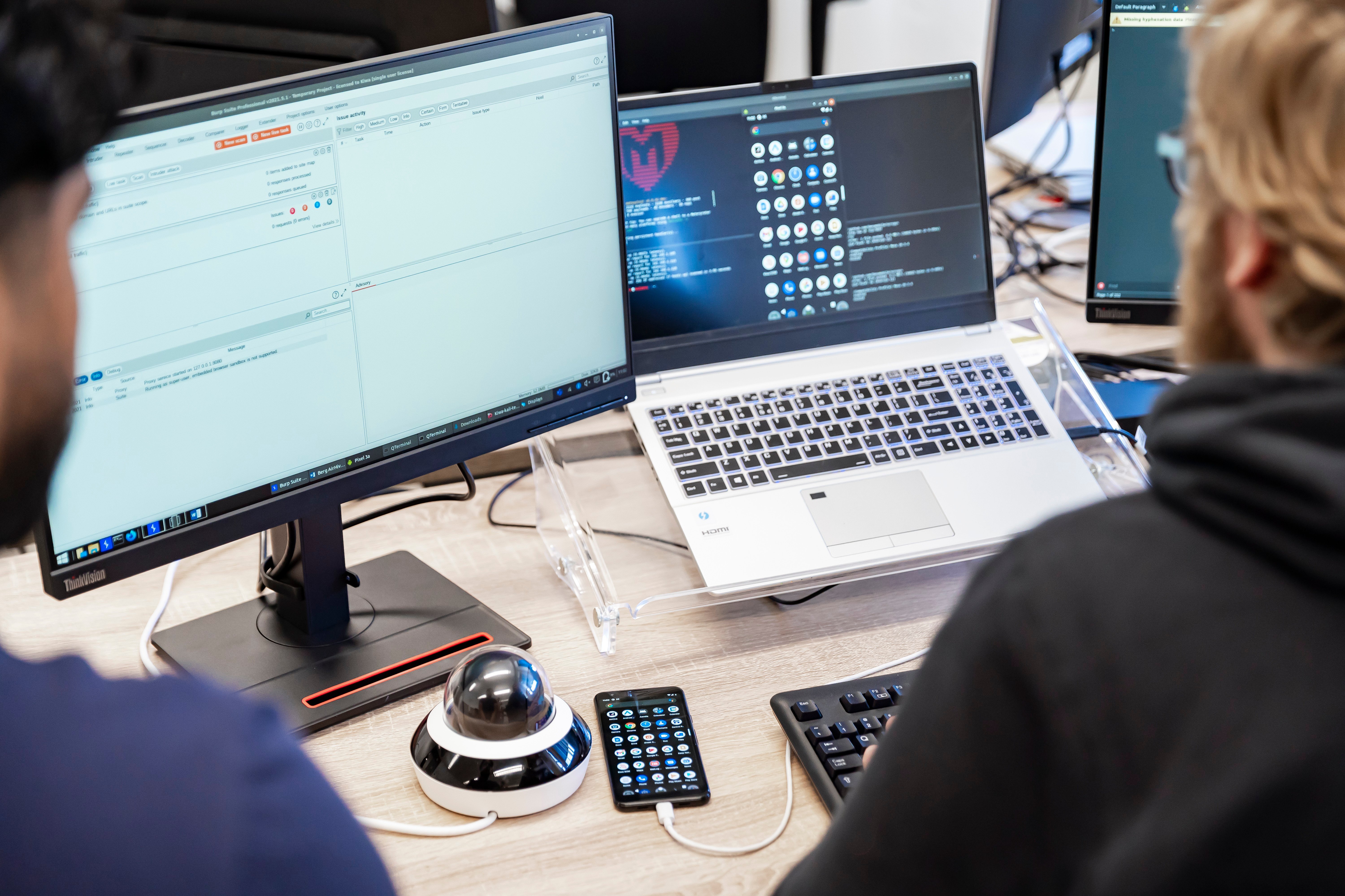 People at a desk with multiple computer monitors, a laptop, and a smartphone displaying software interfaces and app icons.