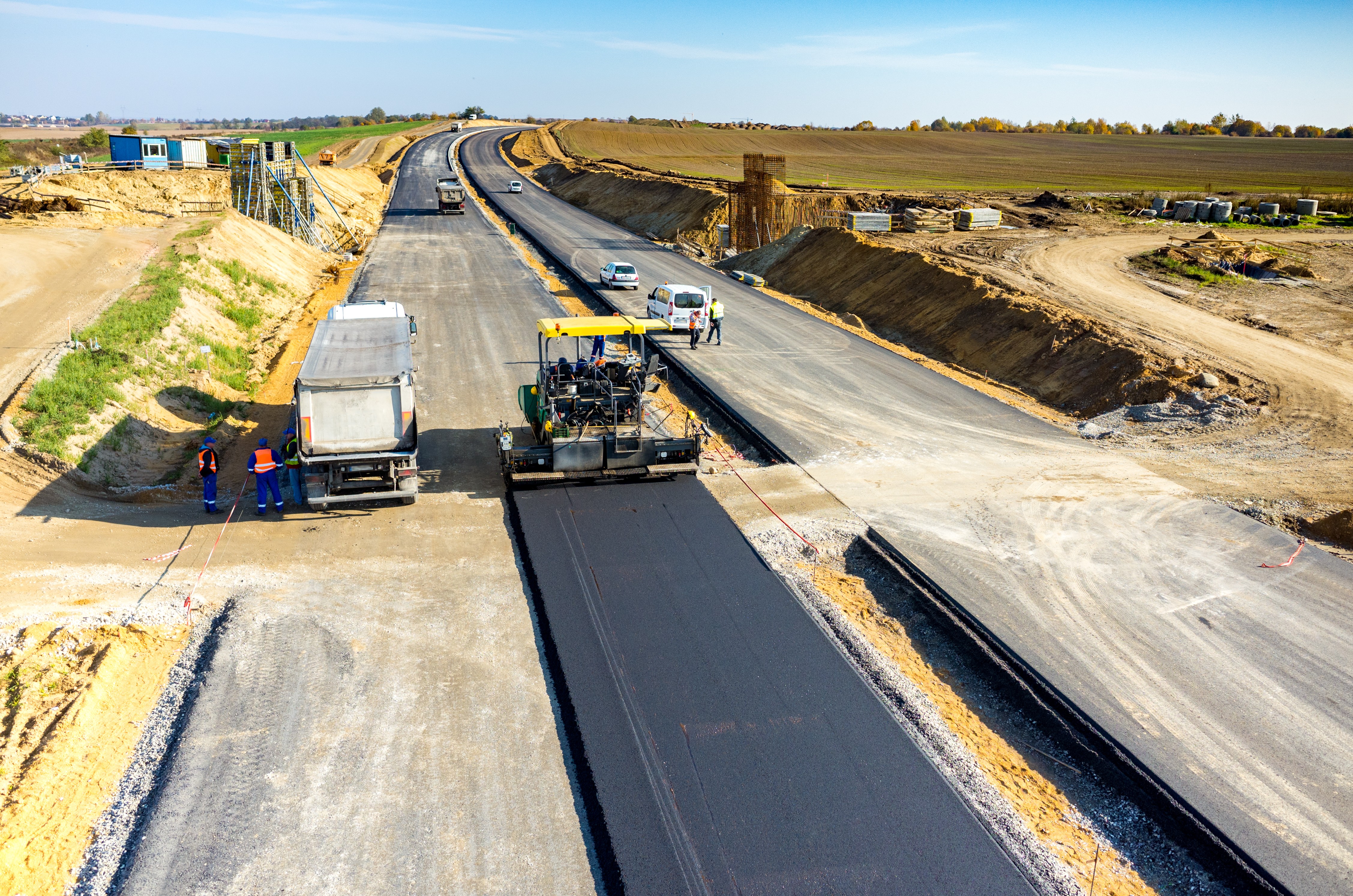 Paving machine applying new Kiwa tested asphalt on a highway under construction, surrounded by construction workers and vehicles, in a rural area