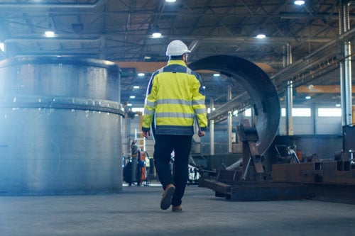 Statybos pramonės specialistas su apsauginiu šalmu ir ryškia geltona liemene vaikšto gamybos ceche tarp didelių metalinių konstrukcijų – Kiwa produktų ir procesų sertifikavimas statybos sektoriuje
