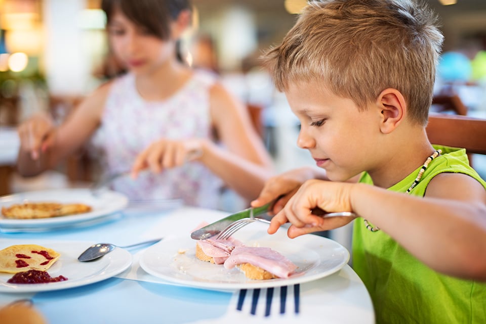 Niño comiendo en un restaurante, con platos de comida como pan y jamón sobre la mesa.