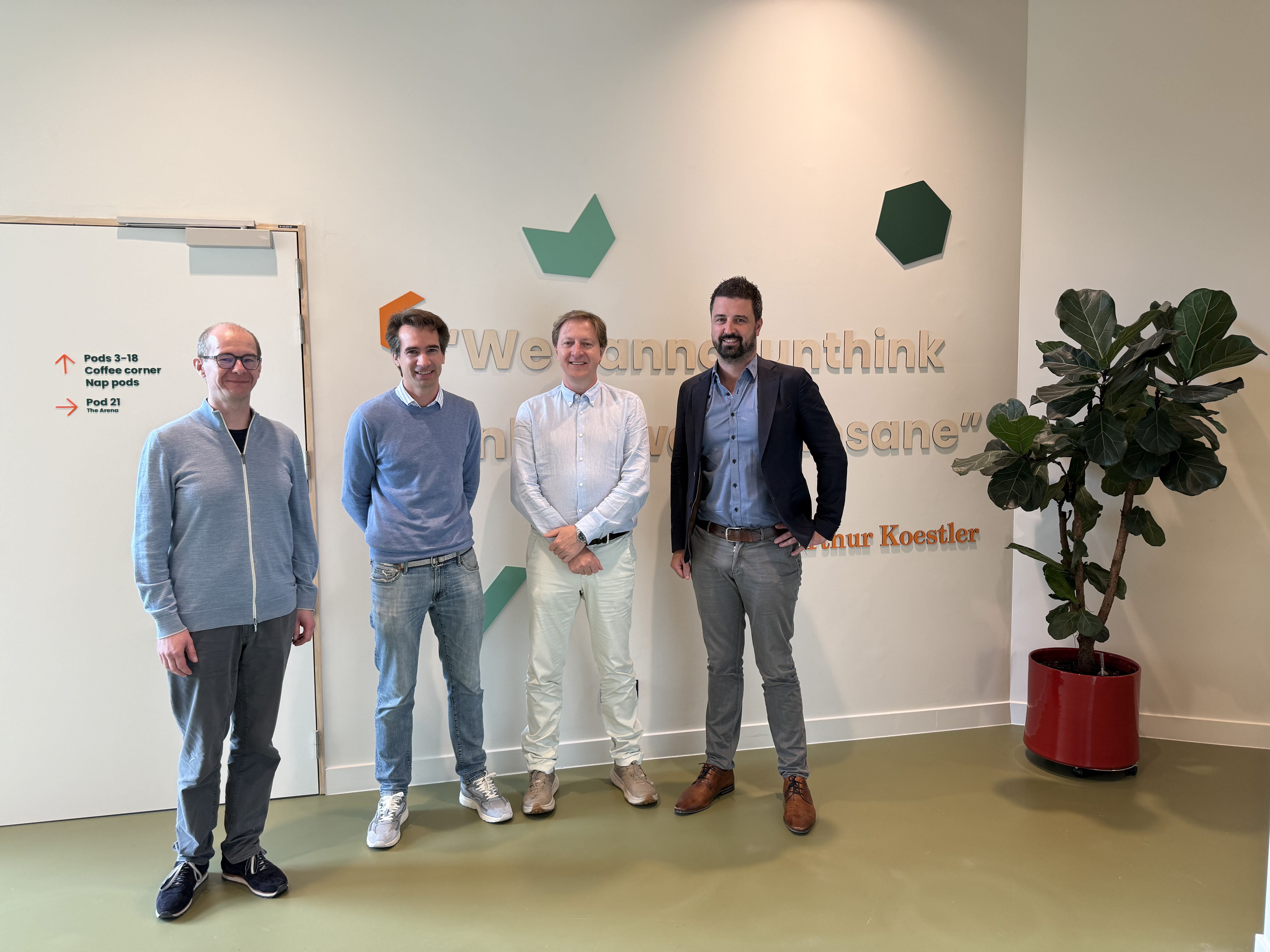 Four men standing in an office lobby with wall art, a potted plant, and a whiteboard sign.