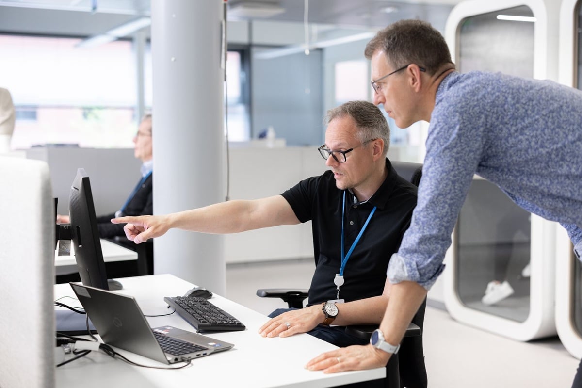 Two men in an office discussing a computer screen, with laptops, keyboard and workstations in a modern workspace.