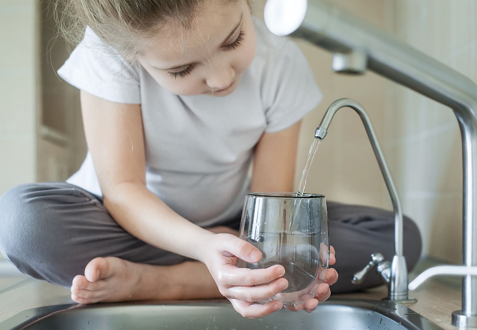 Young girl filling a glass with tap water at the kitchen sink, holding the glass under running water.