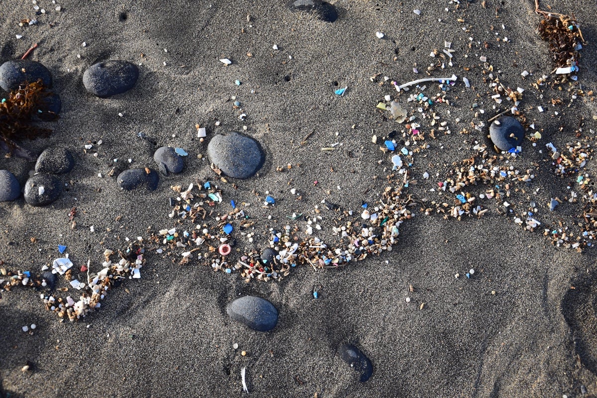 Zwart zandstrand met verspreide keien en een strook aangespoeld plasticafval en microplastics.