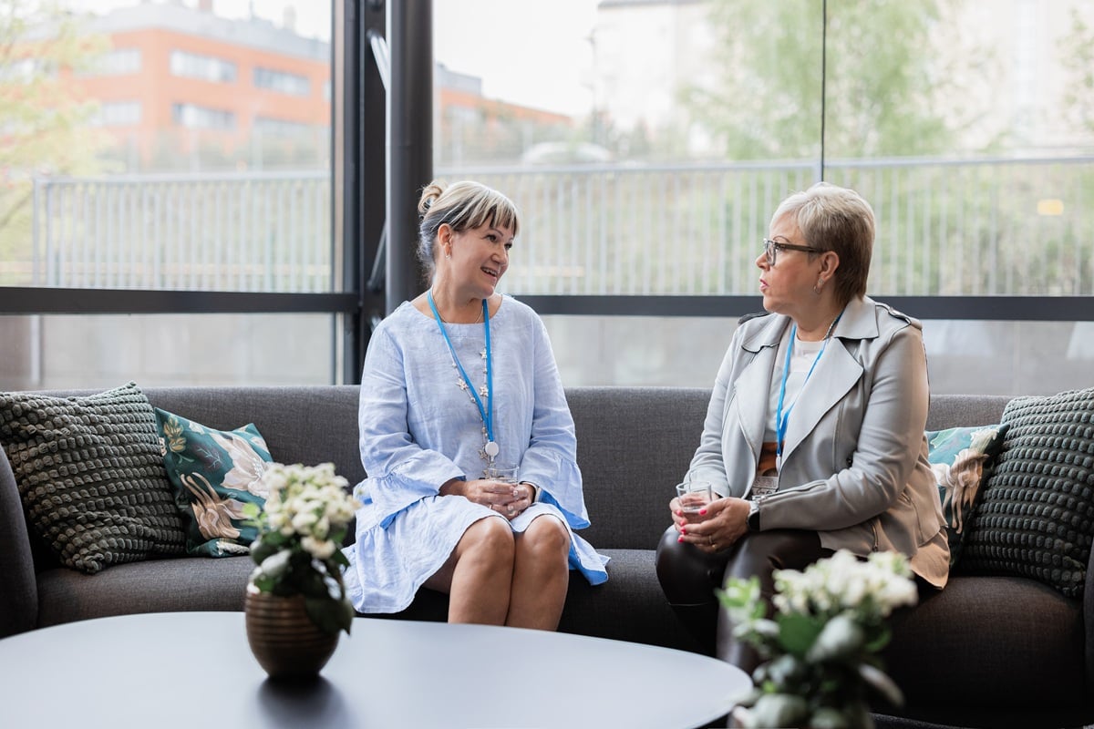 Twee vrouwen zitten en praten op een bank in een lichte lounge. Op de voorgrond staan tafels met bloemen en kussens op de bank.