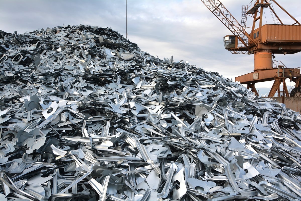 Grote stapel schrootmetaal op een industrieterrein naast een havenkraan.