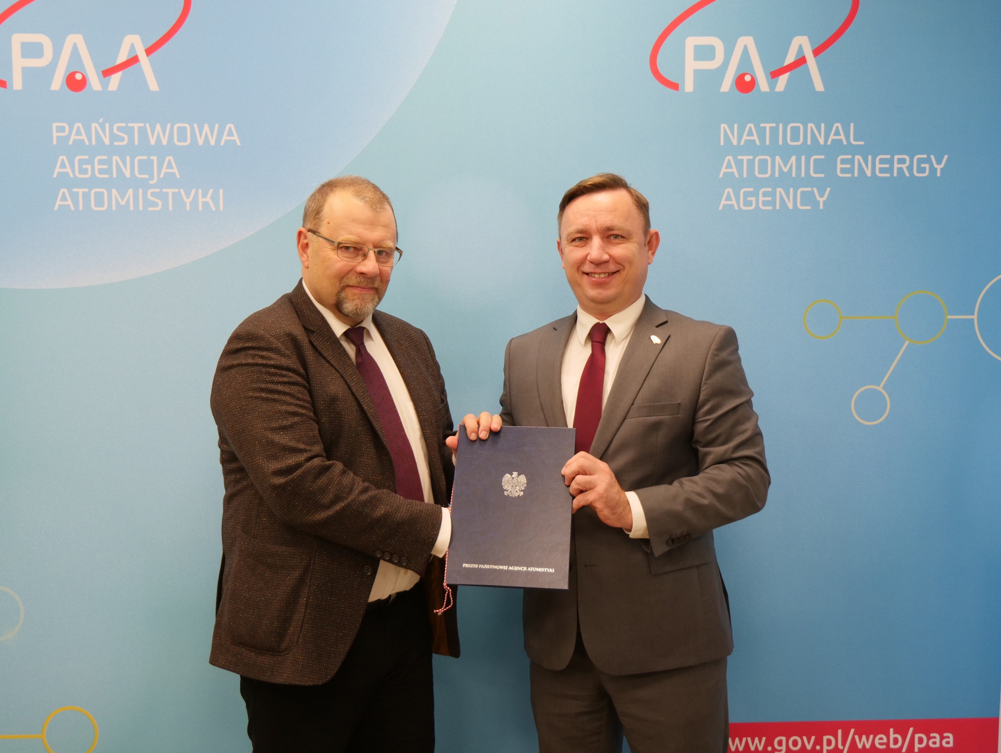 Dwóch mężczyzn w garniturach stoi przed niebieskim tłem z logo Państwowej Agencji Atomistyki, trzymając dokument z polskim godłem.