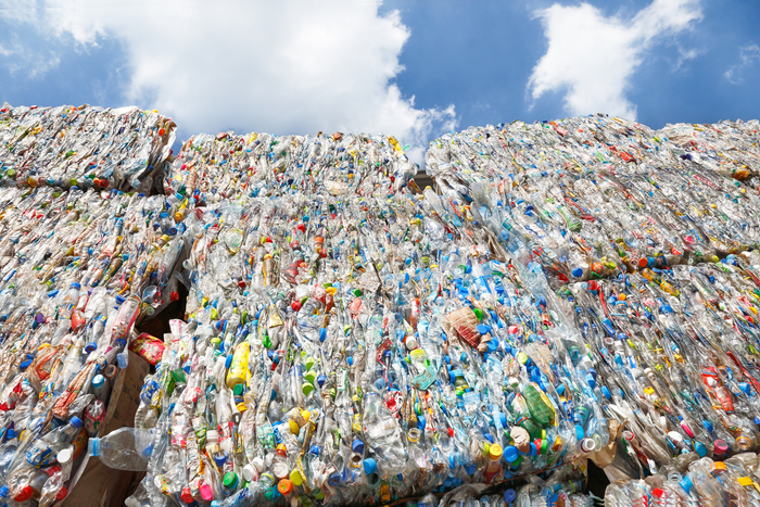 A large stack of compressed plastic bottles bundled for recycling is shown against a bright blue sky