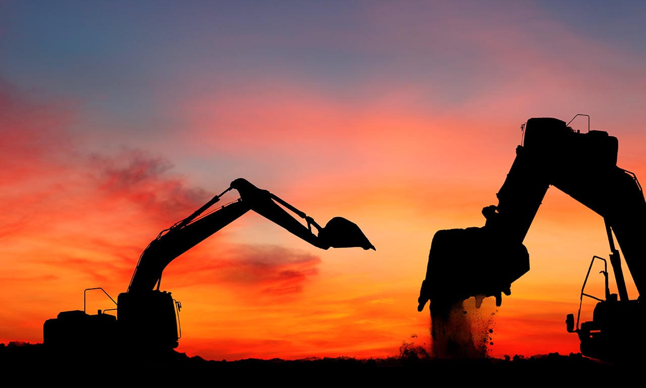 Siluett av en gr&auml;vmaskin i r&ouml;d-gul-orange skymning