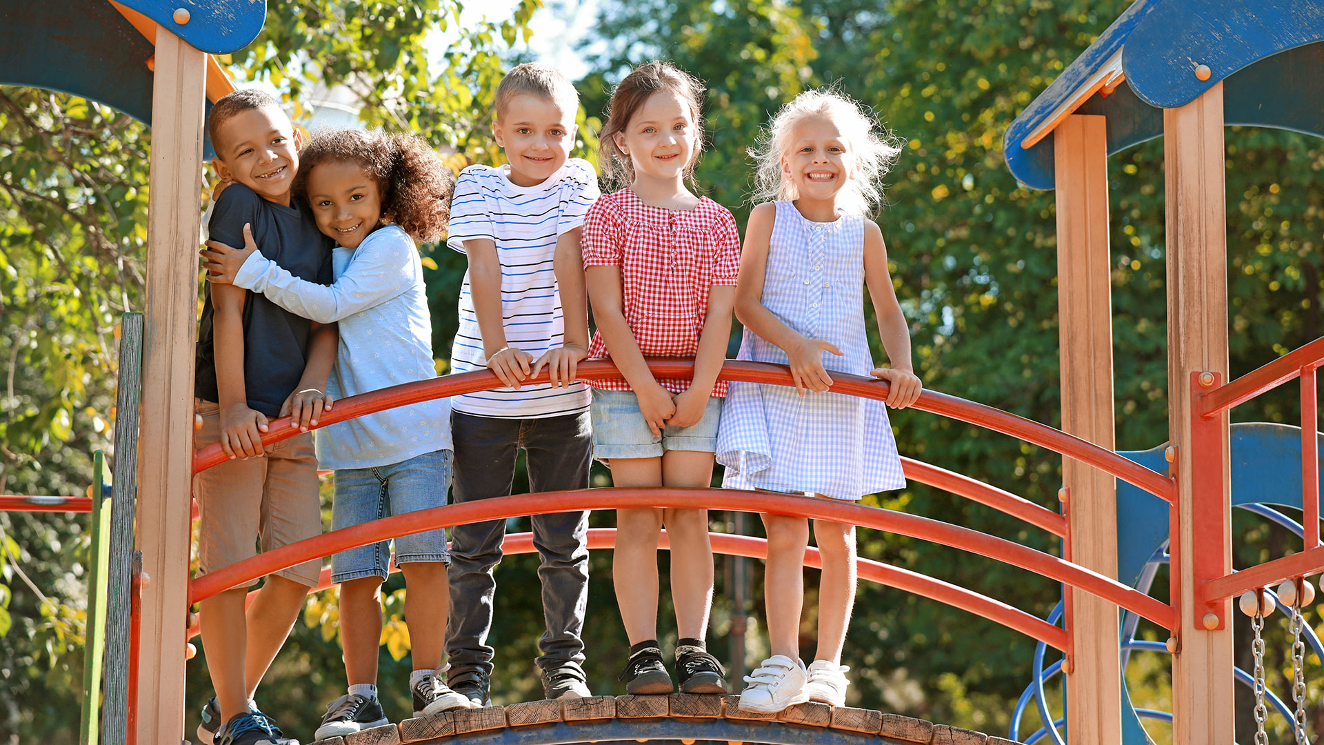 Glada somriga barn står uppställda på rad på en liten gångbro i en lekpark. 