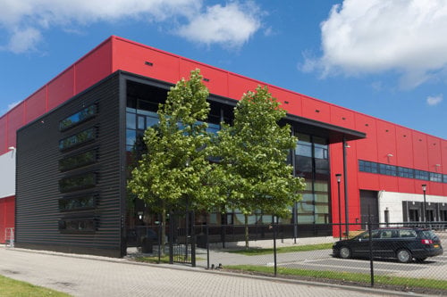 Stor röd och svart industribyggnad med stora fönster, träd framför entrén.