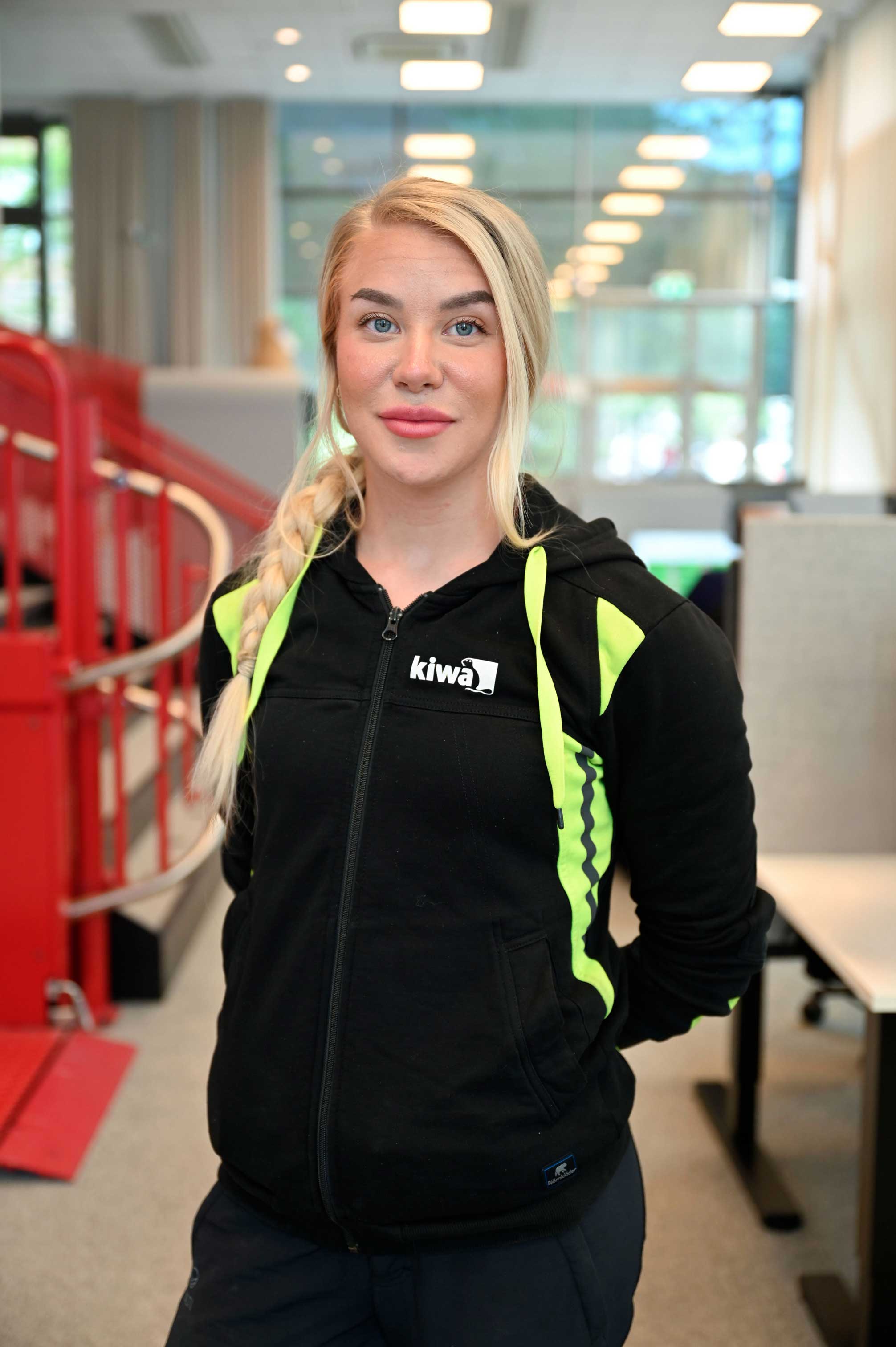 Porträtt av en kvinna i svart hoodie med neongula detaljer och logotypen Kiwa, stående inomhus.