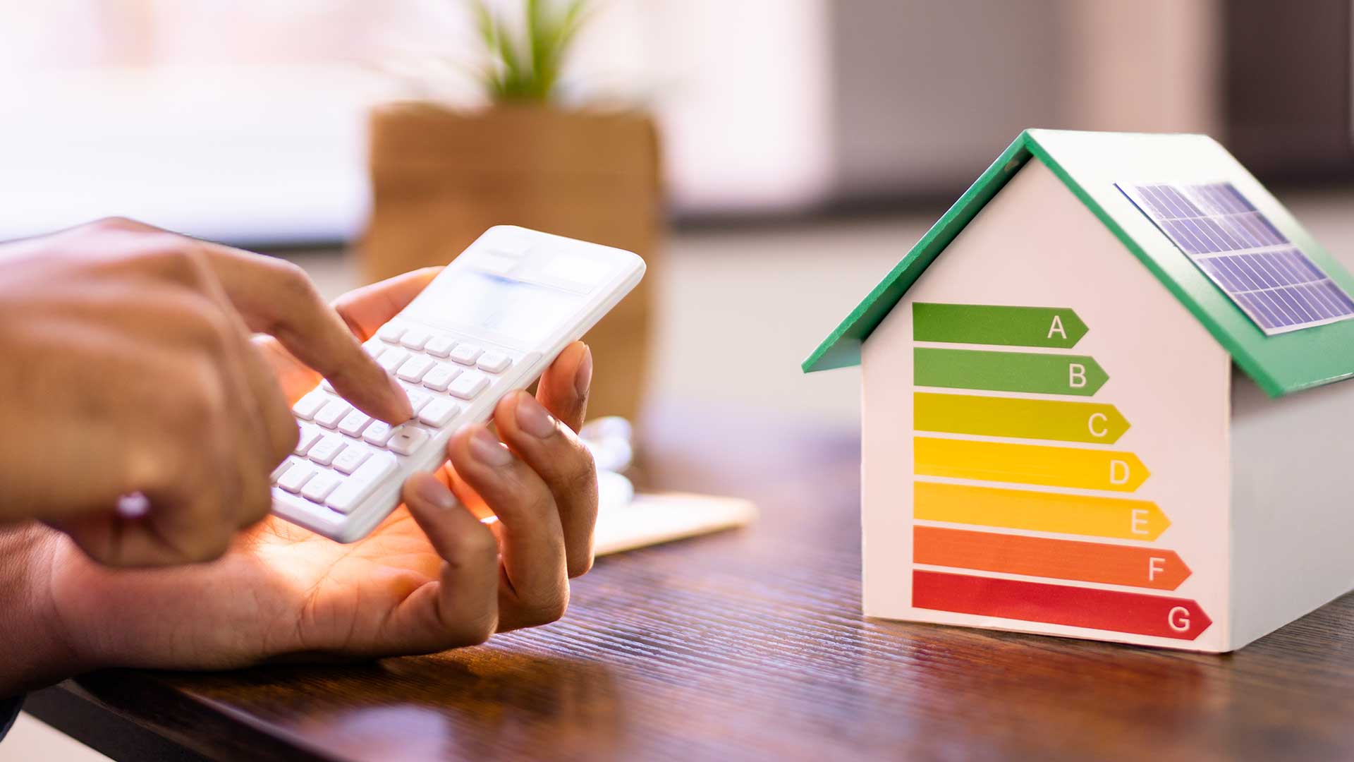 Person använder en miniräknare vid ett bord bredvid en modell av ett energieffektivt hus med solpaneler och energiklassning A–G, som ska symbolisera EU:s energiprestandadirektiv (EPBD) och svenska regelkrav om energideklaration för byggnader.