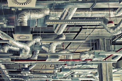 Ventilationssystem uppe i taket med flera rör och fläktar i en industrilokal.