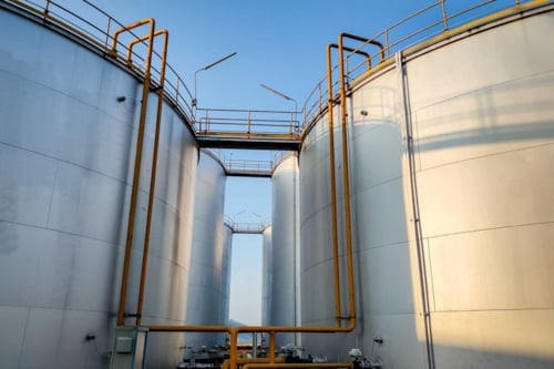 Stora industriella lagringstankar i metall med rör och gångbroar mot blå himmel.