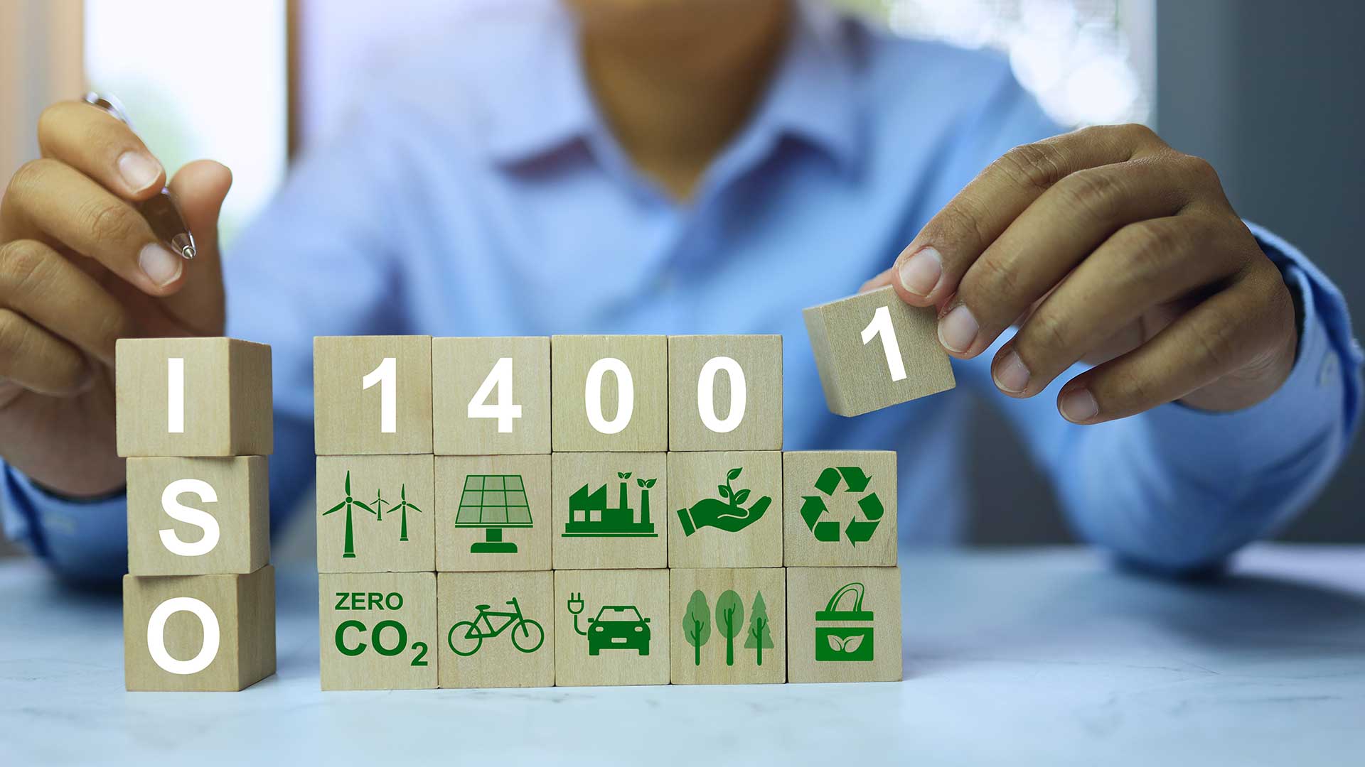 Person som placerar träklossar med ISO 14001 och miljöikoner, symboliserande hållbarhet och miljöledning.