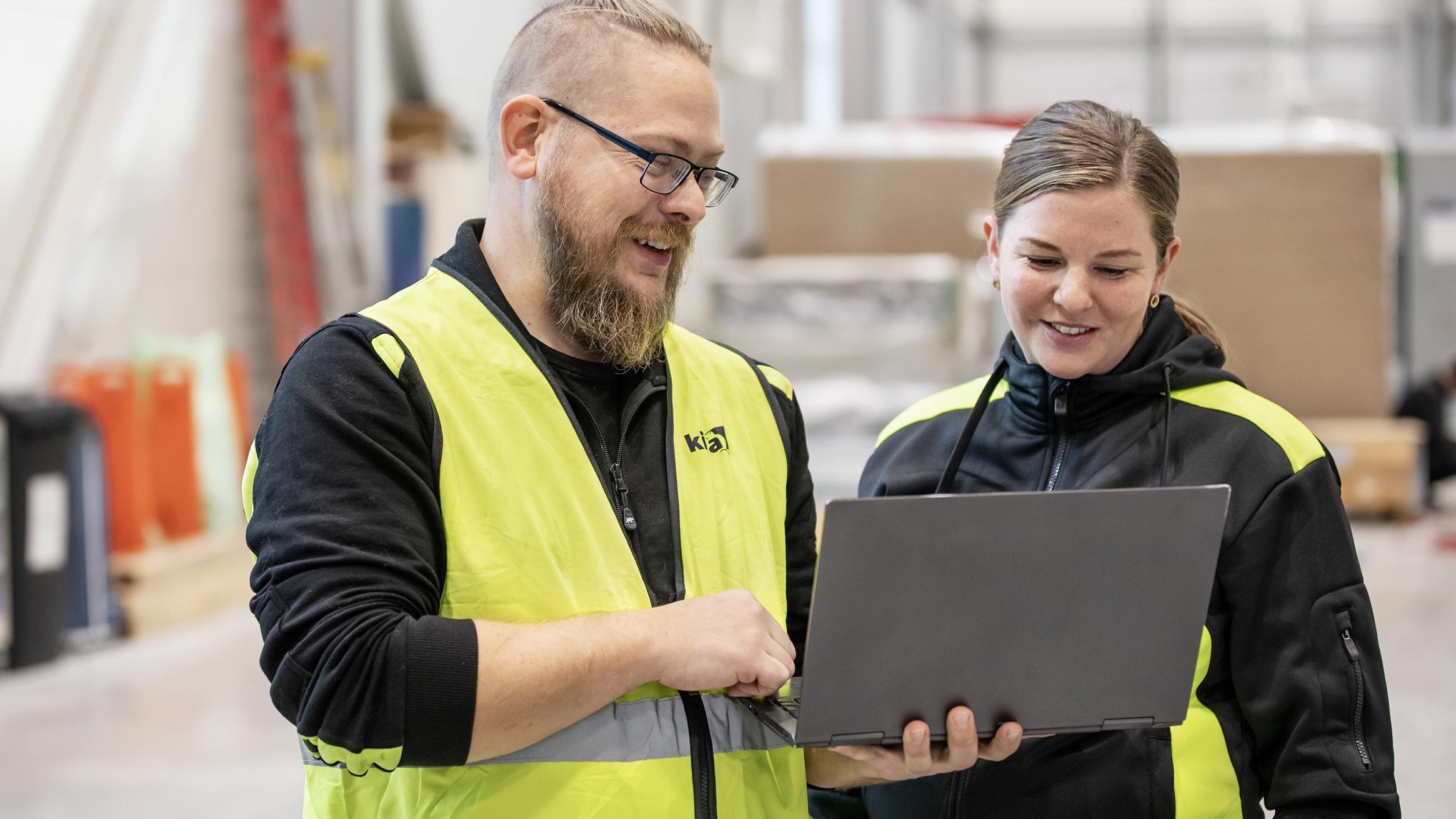 En man och en kvinna i gula varselvästar tittar ner på en laptopskärm som mannen håller i ena handen medan han pekar mot skärmen med den andra. 