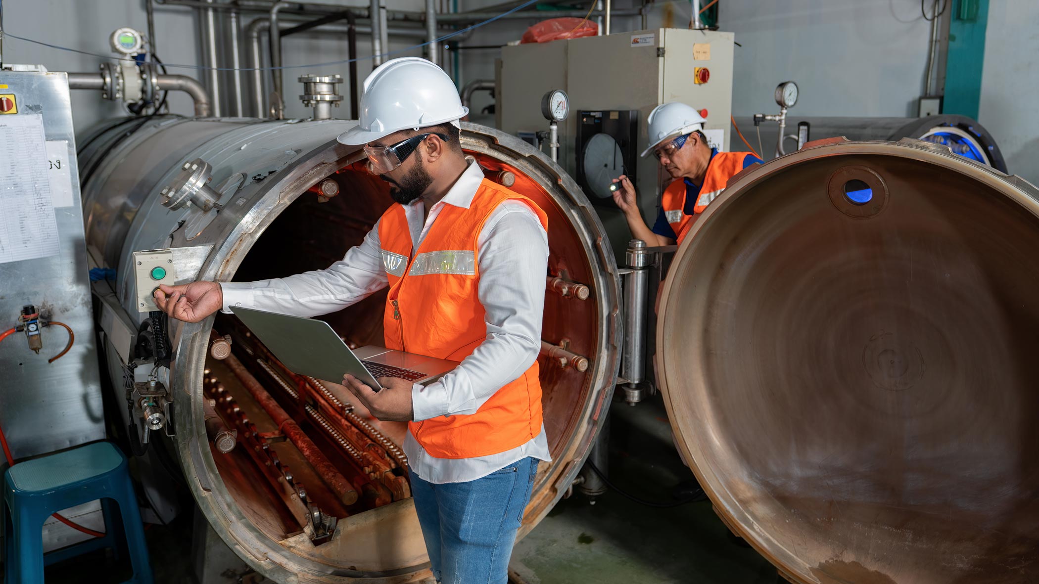 Två tekniker i skyddshjälm och orange varselväst inspekterar en stor industriell trycktank med öppen lucka, varav en använder en laptop.