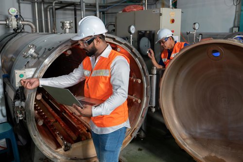 Två tekniker i skyddshjälm och orange varselväst inspekterar en stor industriell trycktank med öppen lucka, varav en använder en laptop.