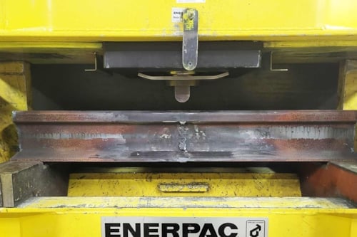 Gul hydraulisk press som böjer en räl i stål, så kallad böjprovning av räl som en metod inom förstörande provning, för att verifiera att rälen uppfyller ställda hållfasthetskrav.