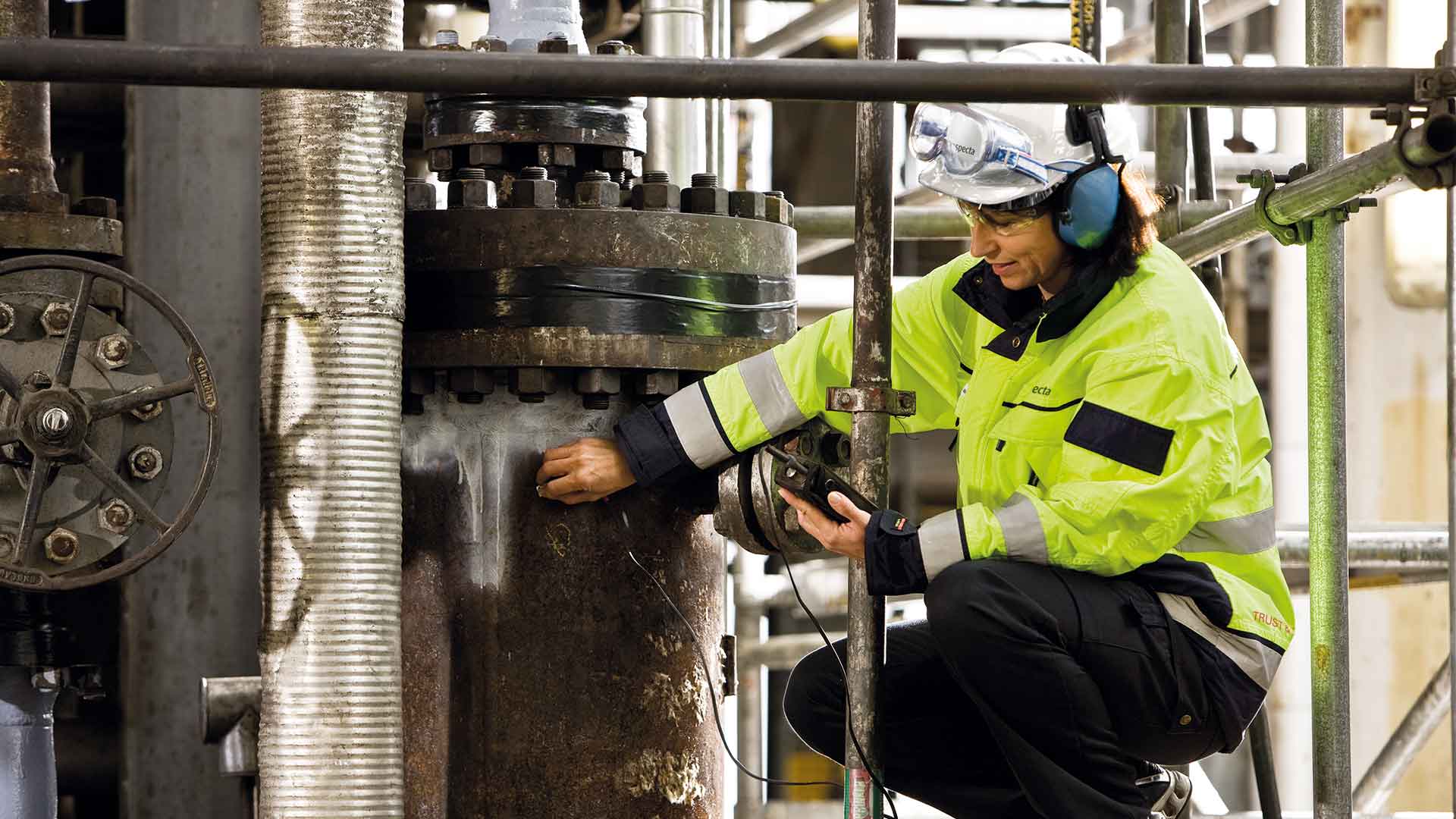 Person i fluorescerande skyddskläder och hörselkåpor som utför ultraljudsprovning i en industrimiljö.