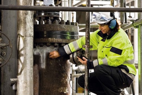 Person i fluorescerande skyddskläder och hörselkåpor som utför ultraljudsprovning i en industrimiljö.