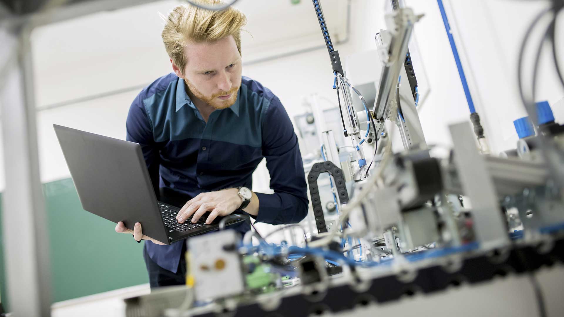 En man i mörkblå skjorta använder en laptop vid industriell utrustning i en verkstad.