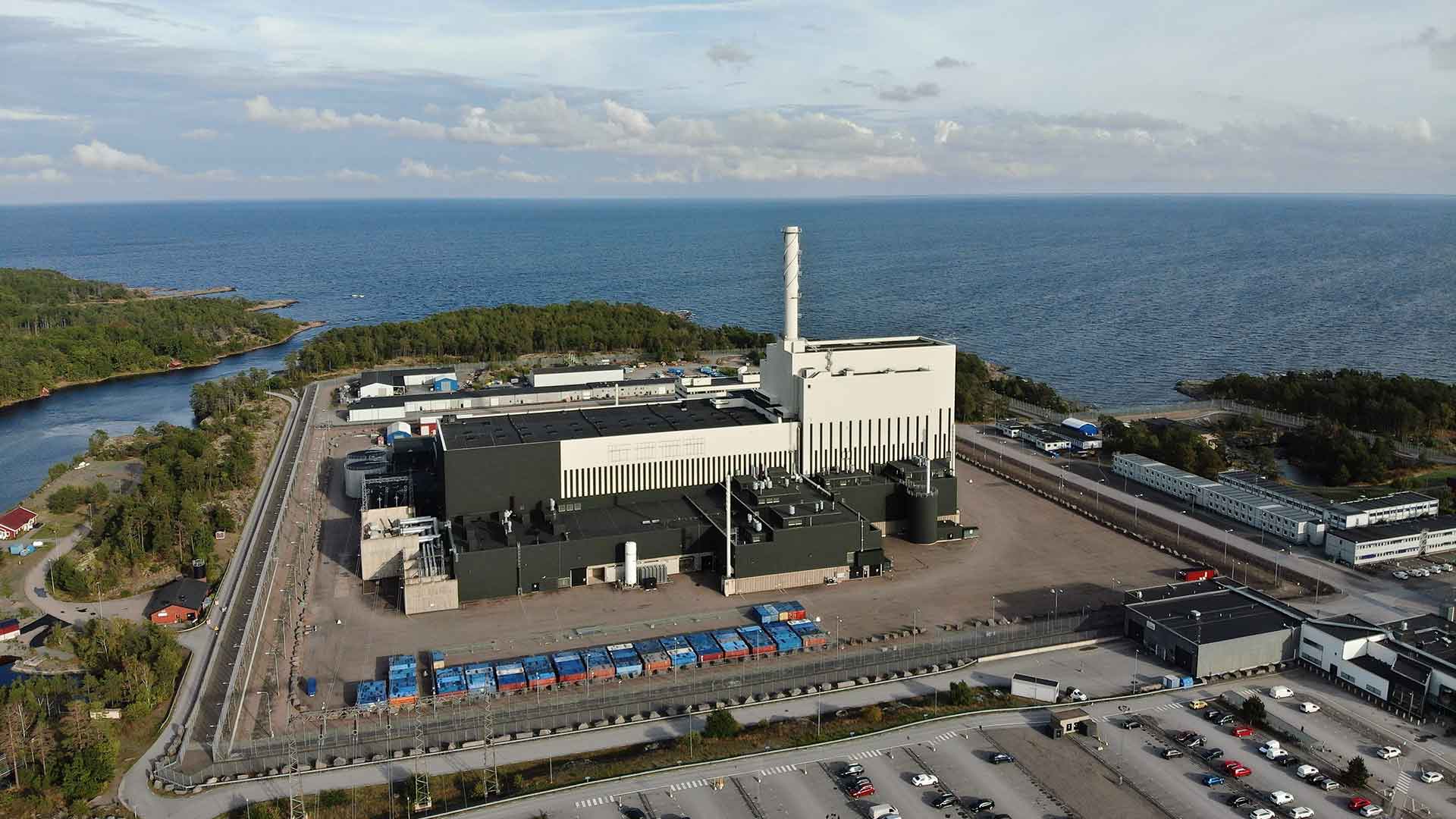 Flygvy över Oskarshamns kärnkraftverk vid kusten, omgivet av hav, vägar och parkeringar.
