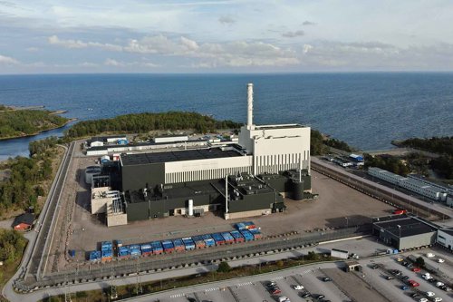 Flygvy över Oskarshamns kärnkraftverk vid kusten, omgivet av hav, vägar och parkeringar.