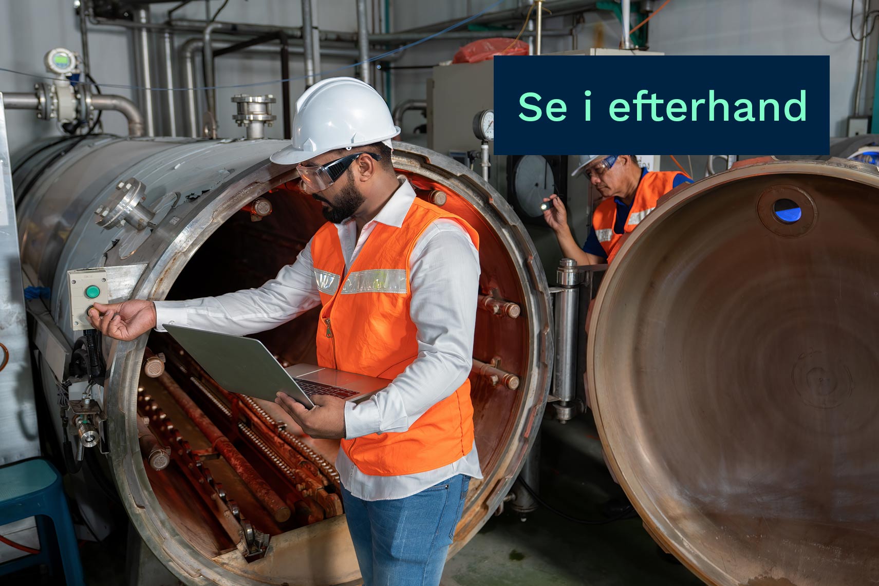 Två industriarbetare i skyddsutrustning inspekterar en stor trycktank med laptop i en fabrik.