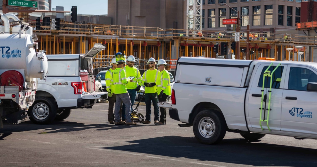 T2UE engineers in bright safety gear gather near utility trucks at an urban job site, with a building under construction in the background