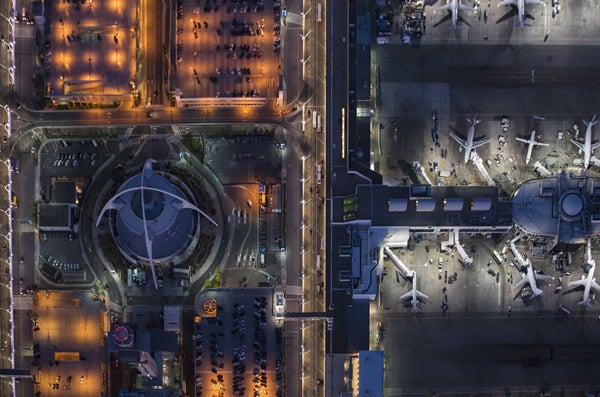 Aerial view of a busy airport terminal of LAX at night, with multiple airplanes parked at the gates and a brightly lit central structure surrounded by parking areas and roads.