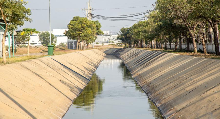 Concrete drainage canal lined with trees on a sunny day, featuring industrial buildings and power lines in the background