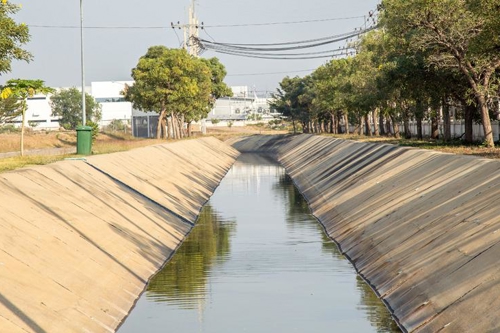 Concrete drainage canal lined with trees on a sunny day, featuring industrial buildings and power lines in the background