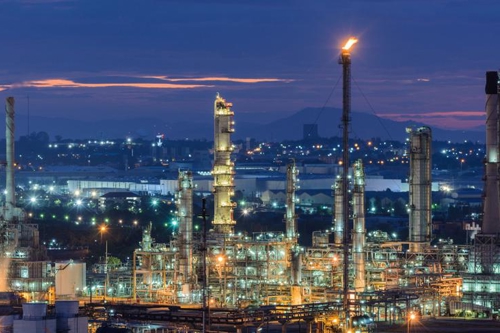 Illuminated oil refinery at dusk with industrial towers and a backdrop of a glowing cityscape