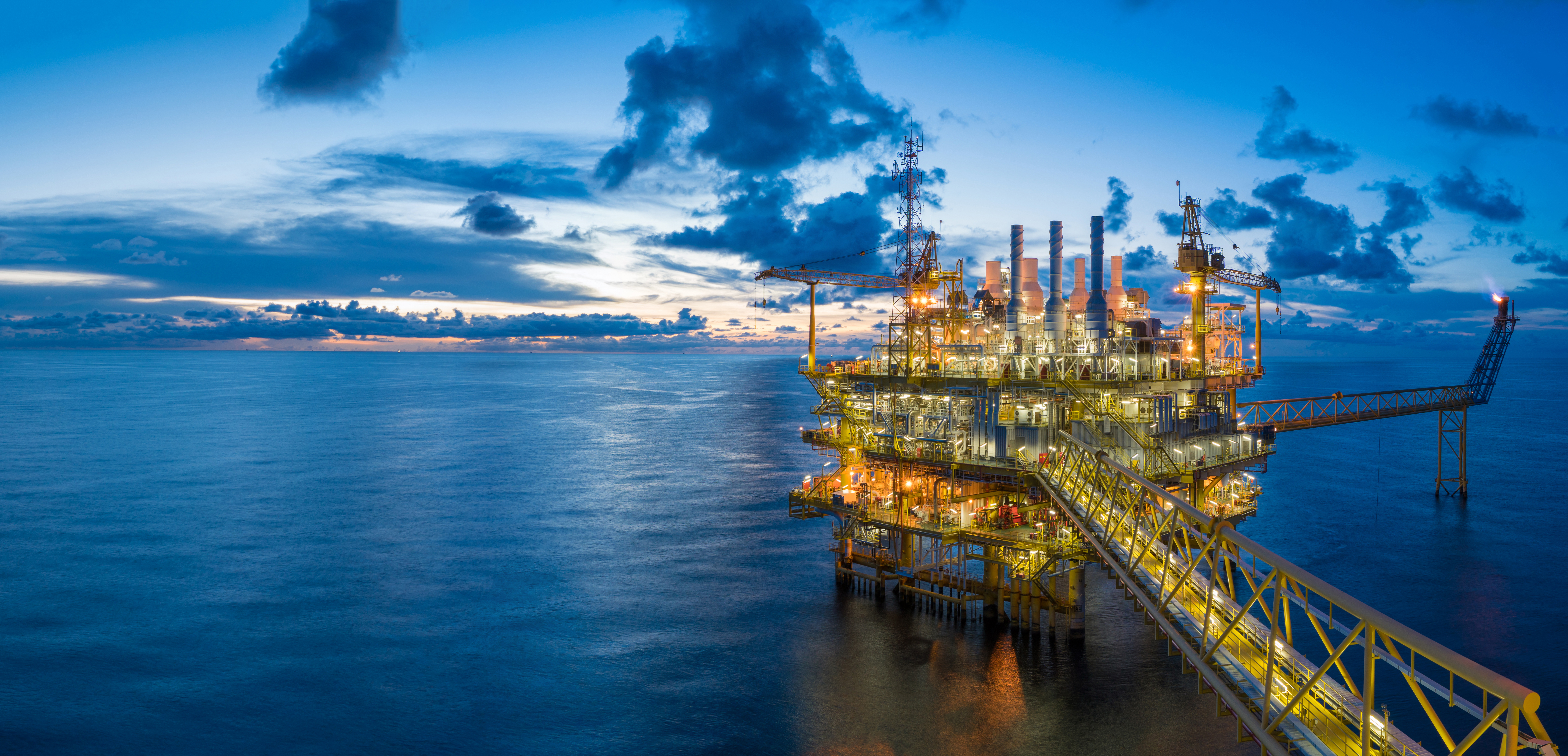 Offshore oil rig at sunset with illuminated structures against a blue ocean and cloudy sky background.